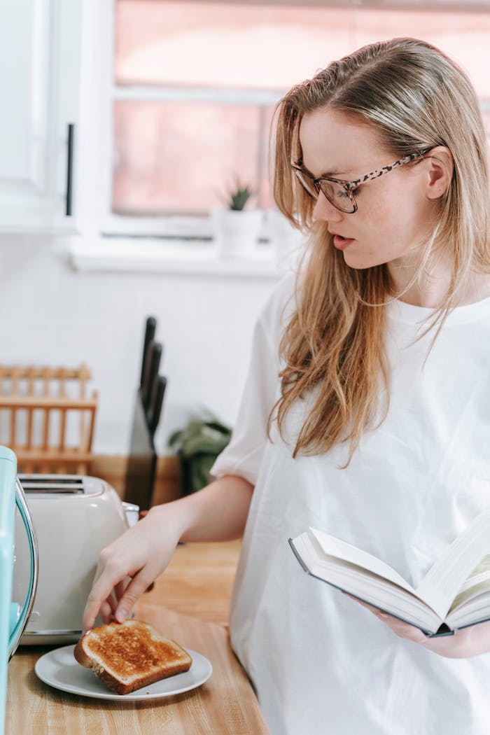Young woman reading a book while preparing breakfast toast in a modern kitchen setting.